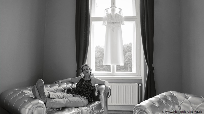 Hochzeitsfotograf Eventschloss Schönfeld - Braut liegt entspannt auf Sofa, kleid hängt dahinter am Fenster