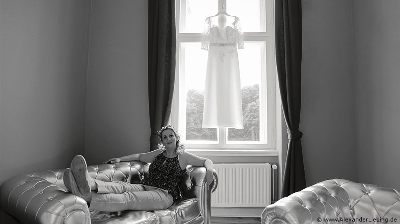Hochzeit - letzte Vorbereitungen und getting ready Hochzeitsfotograf Eventschloss Schönfeld - Braut liegt entspannt auf Sofa, kleid hängt dahinter am Fenster