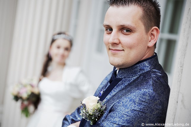 Hochzeitsfotograf Standesamt Magdeburg / Elbelandhaus - Ehemann verschränkt Arme, Braut im Hintergrund