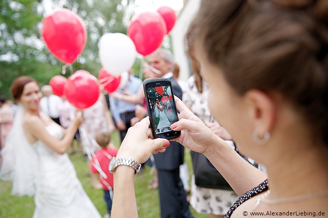 Hochzeit - Hochzeitsfeier Hochzeitsfotograf Standesamt Barleben / Robinien-Hof Gommern - ein Gast fotografiert die Braut, die gerade Luftballons an die Gäste verteilt